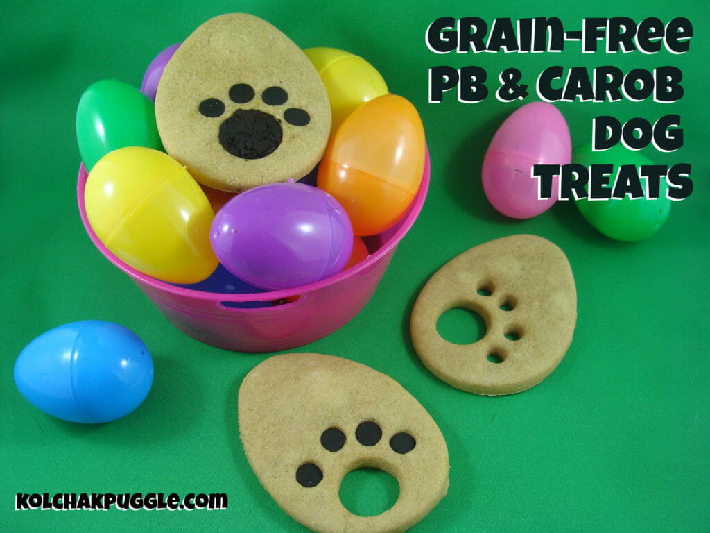 A bowl of plastic coloured easter eggs is shown with Easter peanut butter and banana dog treats around them. The egg shaped have paw shapes cut into them and they are filled with carob. 