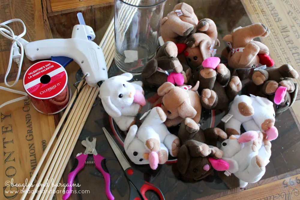 The supplies to make a DIY bouquet made from plush toy dogs lays on the floor. Clockwise from top right: Glue gun, Vase, plush ty dogs, scissors, wood dowels, ribbon.