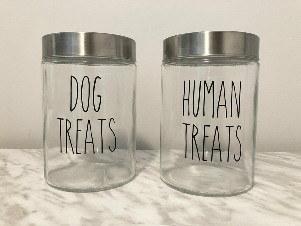 Two DIY clear glass dog treat jars sit on a marble table. They say "dog treats" and "human Treats"