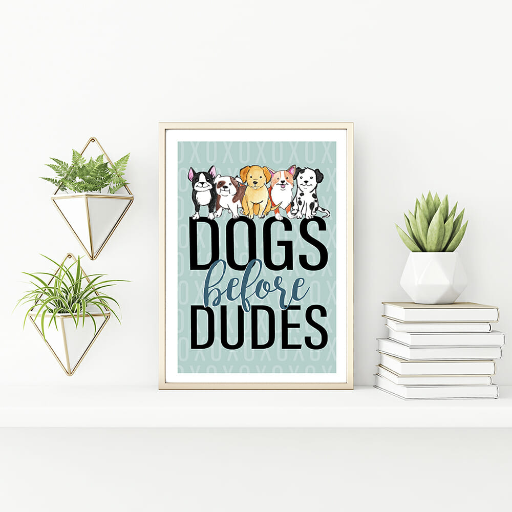 A vertical gold metallic frame sits on a table with plants in pots and a pile of books. In the frame there is a free printable art print with a blue background print and a watercolour boston terrier, an english bulldog, a yellow labrador, a corgi and a dalmation. Text says: Dogs before dudes
