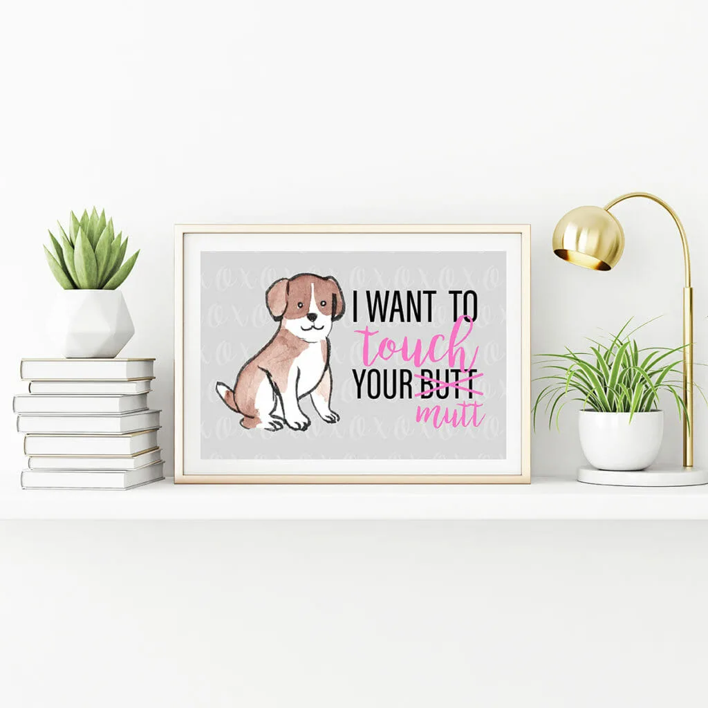 A horizontal gold metal frame standing on the table with plants in pots, lamp and pile of books. Inside the frame there is a free printable art print with a grey background and a brown and white dog of no particular breed. Text says: I want to touch you butt. "Butt" is crossed out and replaced with "mutt"