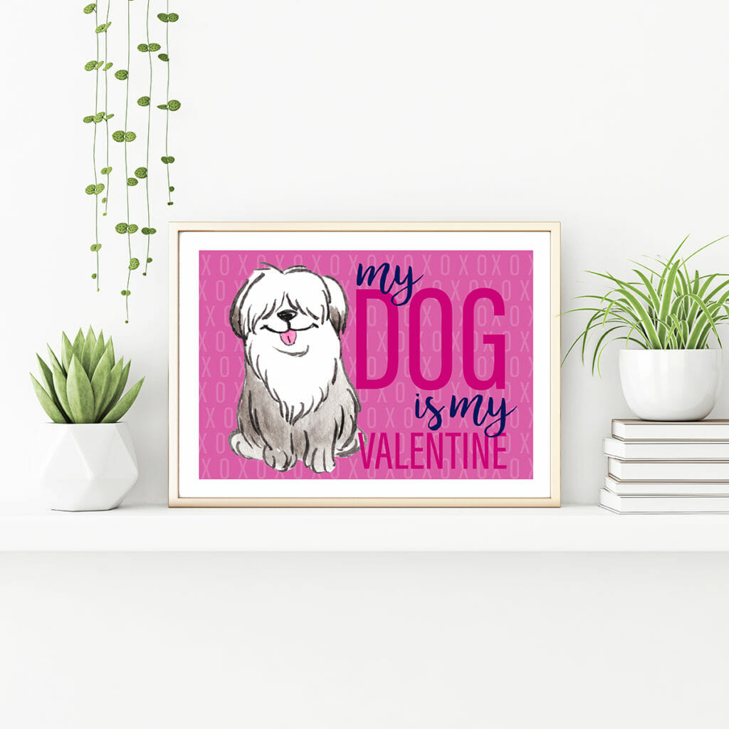 A horizontal gold metal frame standing on the table with plants in pots and pile of books.IN the frame is a free printable art piece with a raspberry coloured background and a watercolor sheepdog. Text says: my dog is my valentine
