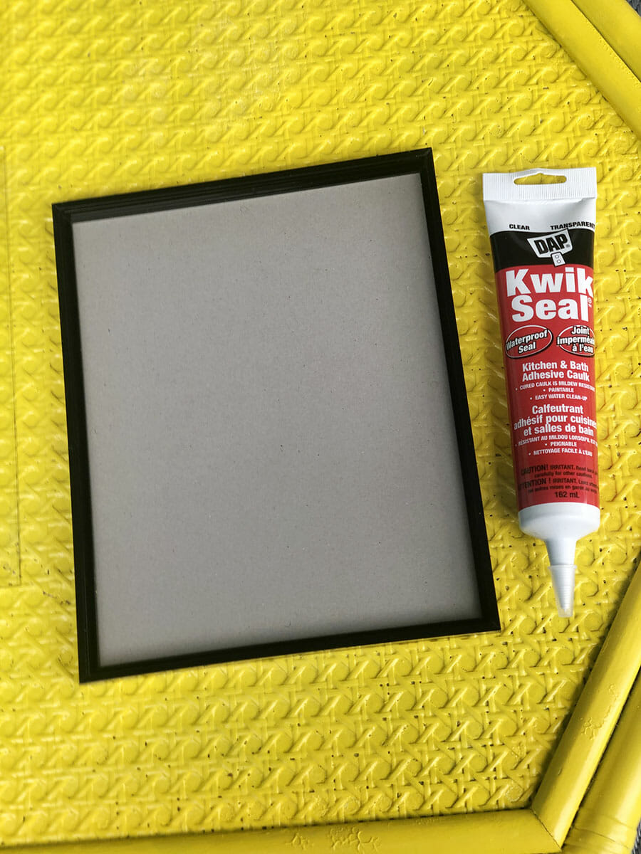 a black frame sits on a bright yellow table with a tube of construction caulking next to it