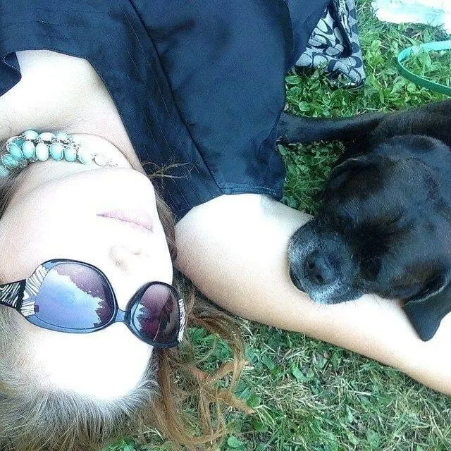 writer Jodi Chick lays in the grass with sunglasses on. Her black puggle Kolchak lays on her arm