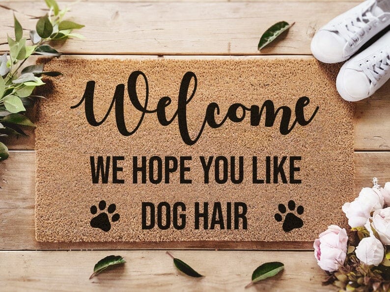 a brown coir dog mat says "welcome. we hope you like dog hair" with shoes and flowers surrounding it.