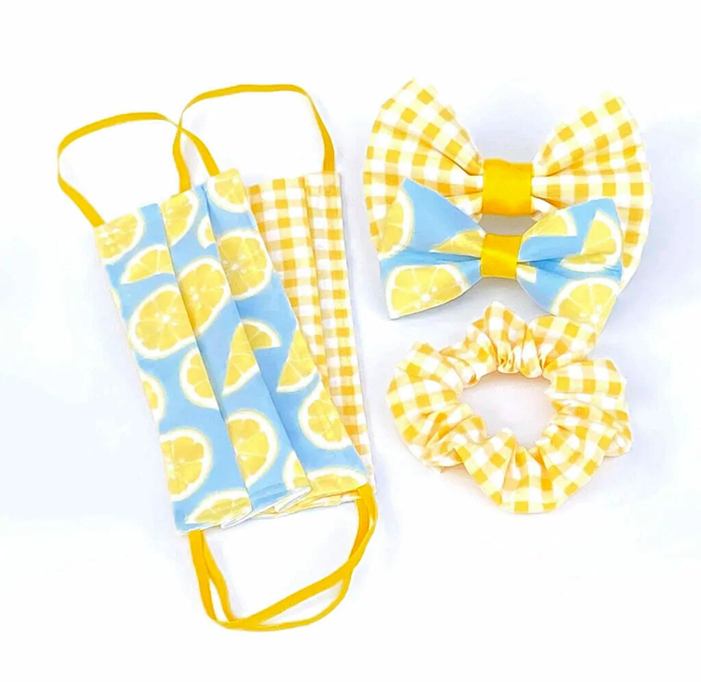 folded, surgical style fabric masks in bright blue, yellow and lemon patterns sit with a matching scrunchie and matching bowties for dogs.