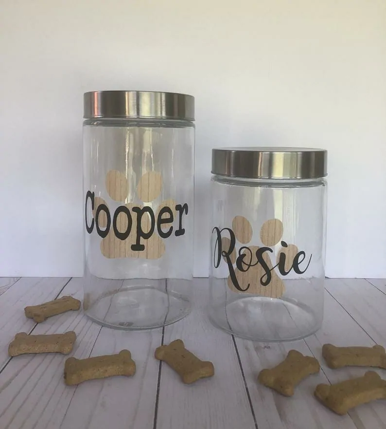 two clear glass dog treat jars with silver lids are on a wood table. They say "Cooper" in type and "Rosie" in script with brown pawprints behind the lettering.