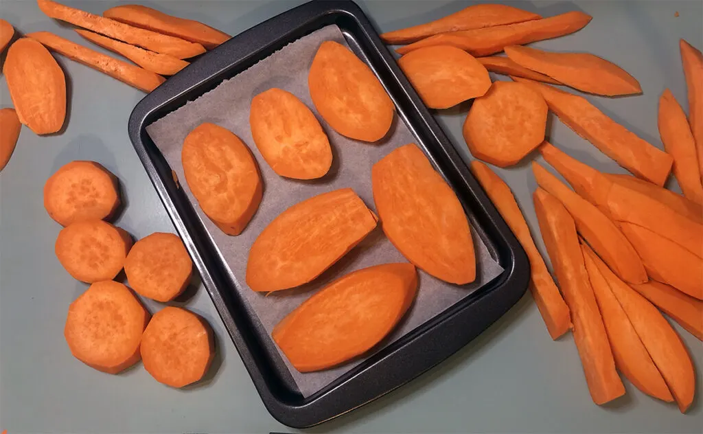 uncooked orange sweet potato slices in various sizes and shapes arranged on and around a baking tray lined with parchment paper
