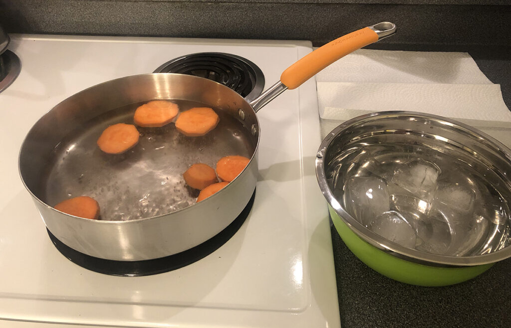 round slices of sweet potato blanching in a pan of boiling water, with an ice bath sitting on the counter next to it