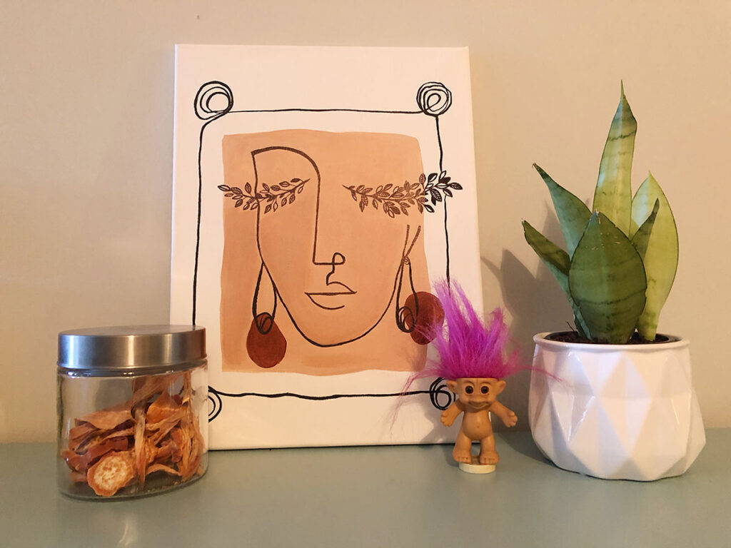 Sweet potato dog chews stored in an airtight glass jar with a painting of a women's face outline on a beige background, a troll doll with pink hair and a houseplant.